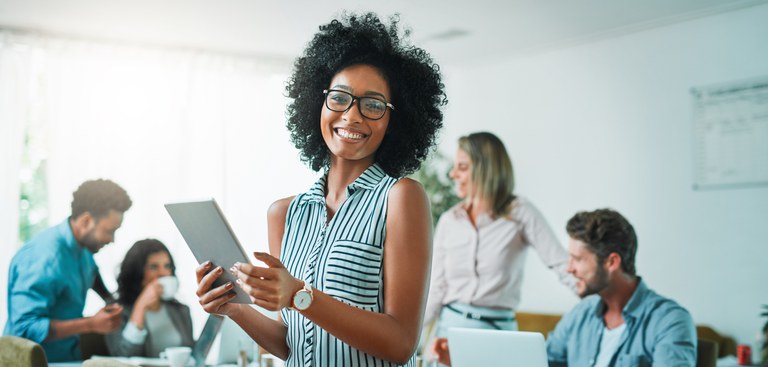 Mulher negra olha para frente e sorri enquanto setgura um tablet. Atrás dela, dois homens e duas mulheres conversam numa mesa com um notebook aberto Mulher negra olha para frente e sorri enquanto setgura um tablet. Atrás dela, dois homens e duas mulheres conversam numa mesa com um notebook aberto