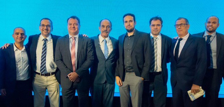 Oito autoridades posam para foto lado a lado, com um fundo completamente azul claro