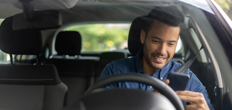 Foto com vista próxima do para-brisa de um carro. Há um homem no banco do motorista que sorri ao utilizar o celular. Foto com vista próxima do para-brisa de um carro. Há um homem no banco do motorista que sorri ao utilizar o celular.