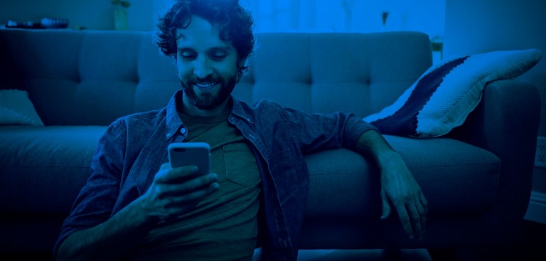 Um homem sorrindo, sentando no chão e encostado no sofá, na sala de casa, olhando para a tela do celular Um homem sorrindo, sentando no chão e encostado no sofá, na sala de casa, olhando para a tela do celular