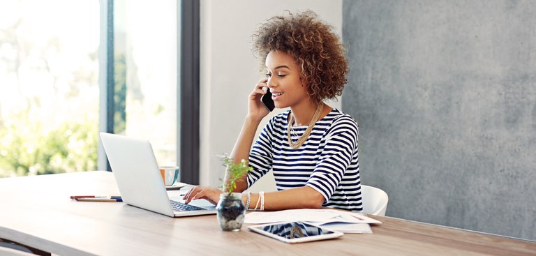 mulher negra atende telefone em frente a notebook, sentada à mesa mulher negra atende telefone em frente a notebook, sentada à mesa