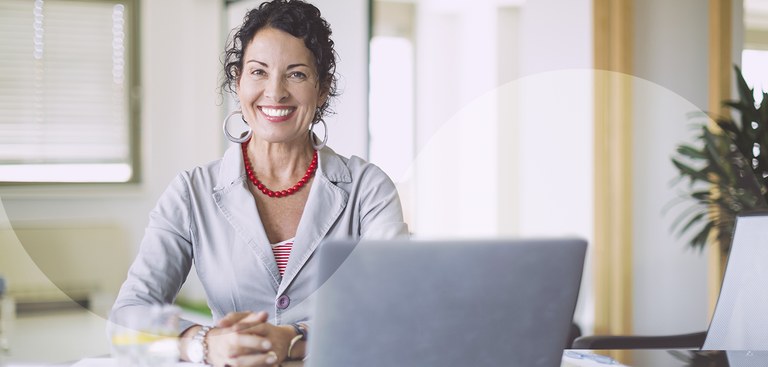 Mulher sorrindo com as mãos apoiadas em uma mesa onde está posicionado um laptop Mulher sorrindo com as mãos apoiadas em uma mesa onde está posicionado um laptop