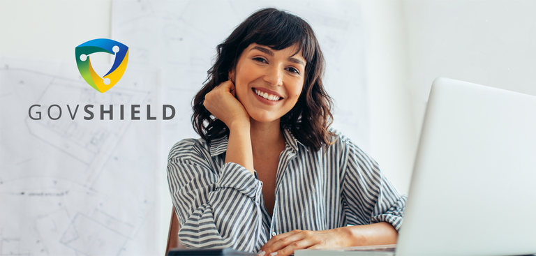 Mulher sorri enquanto sentada à mesa de trabalho com um laptop. No canto superior esquerdo da imagem está aplicada a marca do GovShield Mulher sorri enquanto sentada à mesa de trabalho com um laptop. No canto superior esquerdo da imagem está aplicada a marca do GovShield