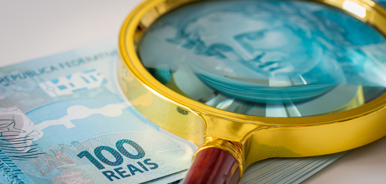 Lupa apoiada sobre um maço de notas de 100 reais Lupa apoiada sobre um maço de notas de 100 reais