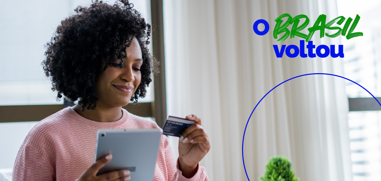 Dentro de casa, uma mulher está com um tablet em uma das das mãos e um cartão de crédito na outra Dentro de casa, uma mulher está com um tablet em uma das das mãos e um cartão de crédito na outra