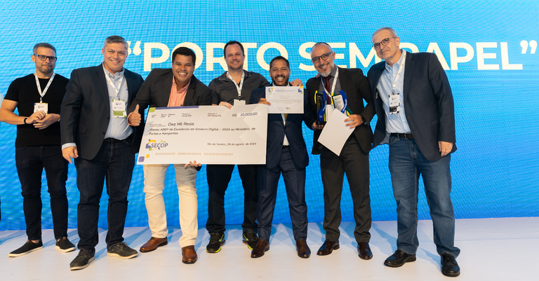 Representantes do Ministério de Portos e Aeroportos, do Serpro e da premiação exibem cheque no palco com fundo azul