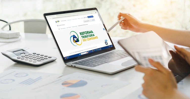 Imagem de um computador sobre uma mesa. Na tela, pode se ver a marca da Reforma Tributária do Conusmo
