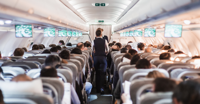 Fotografia do interior de uma aeronave com passageiros nos assentos e aeromoça de uniforme andando pelo corredor