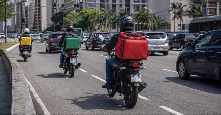 Motociclistas de aplicativo de entrega trafegam por uma avenida movimentada em área urbana, carregando mochilas térmicas coloridas enquanto dividem espaço com carros no trânsito Motociclistas de aplicativo de entrega trafegam por uma avenida movimentada em área urbana, carregando mochilas térmicas coloridas enquanto dividem espaço com carros no trânsito